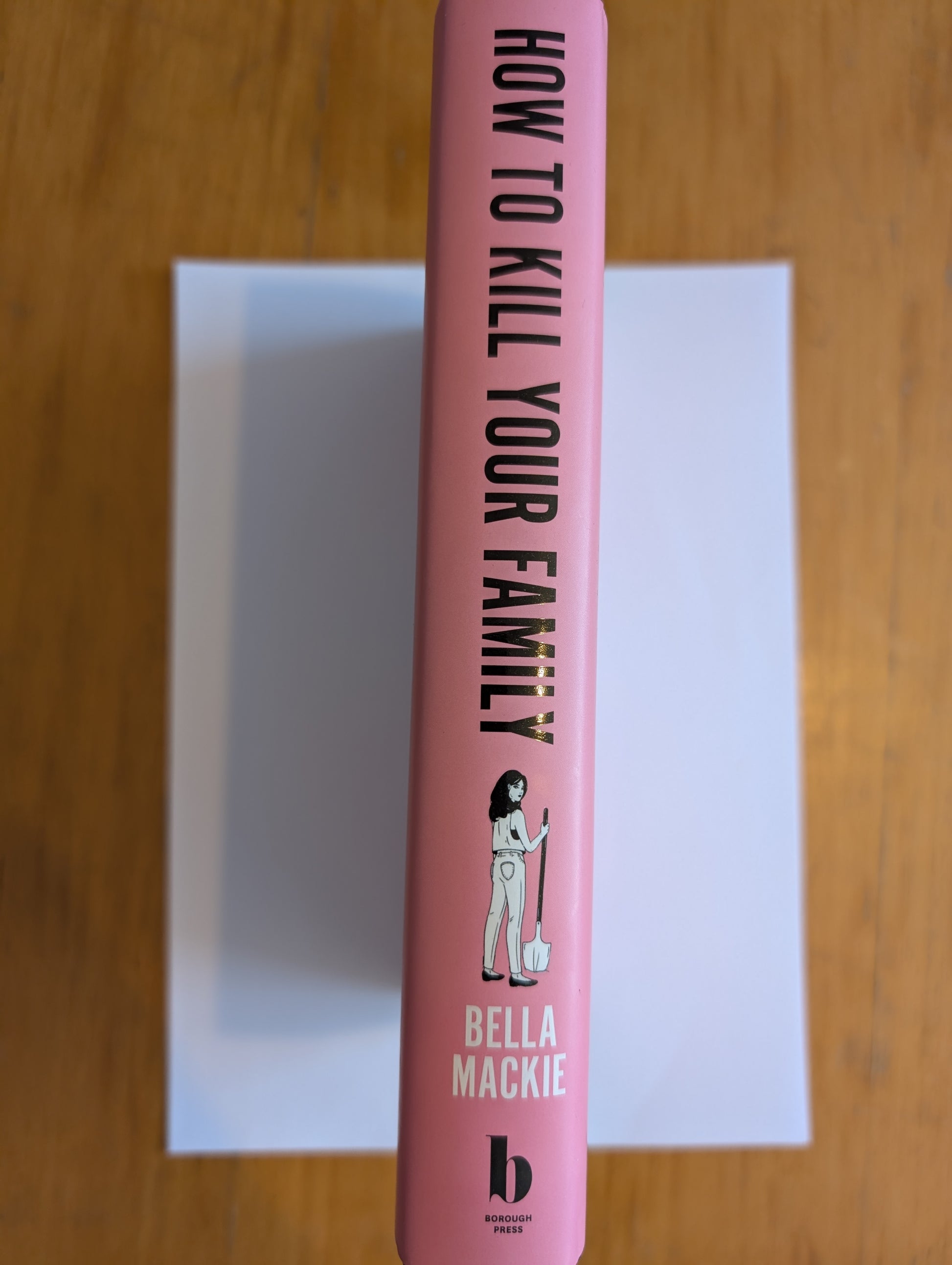 Pink book titled 'How to Kill Your Family' by Bella Mackie on a wooden surface.